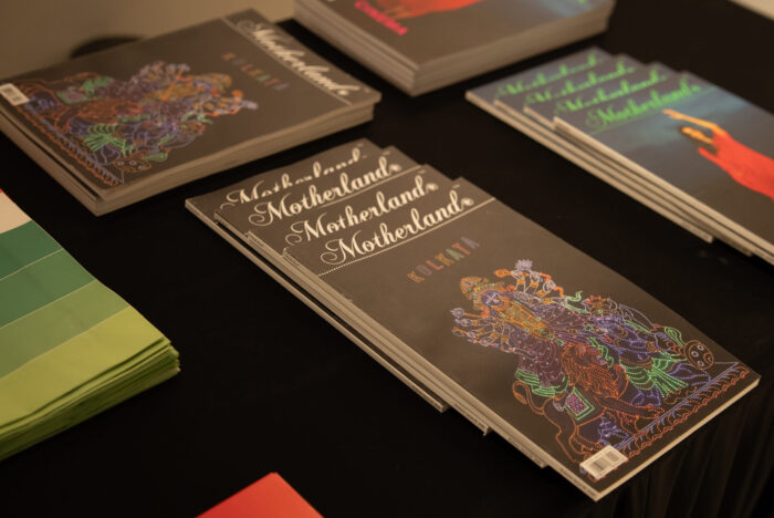 Stack of magazines on a dark table, covers feature ornate, colorful artwork and the word 'Motherland'.