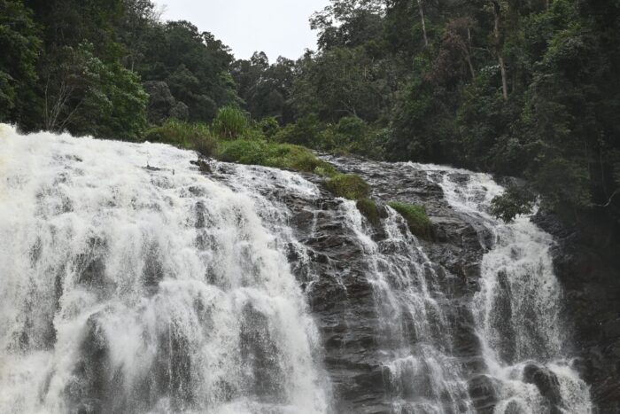 Waterfall rushing over rocky ledges with dense green forest on both sides.
