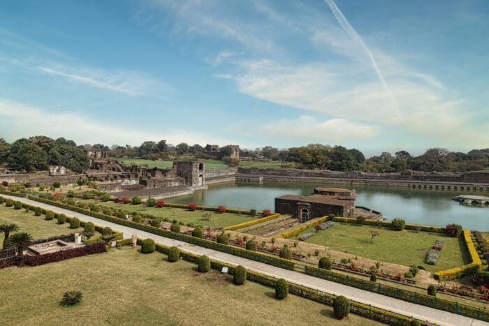 Historic castle ruins set in a formal garden with trimmed hedges, a rectangular moat, and blue sky above.