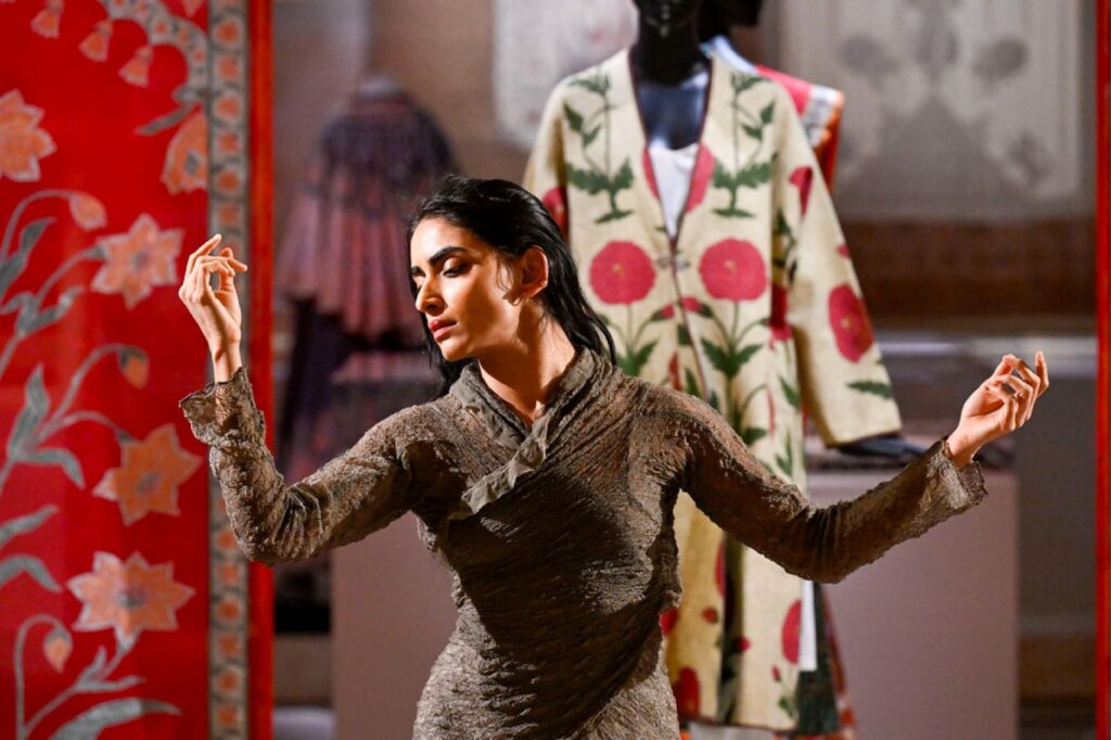 Dancer in a textured brown outfit raises arms in a performance, with a floral-patterned garment on a mannequin in the background.
