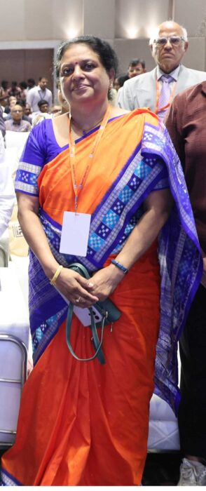 Woman in an orange and purple saree with a lanyard at a conference, hands clasped in front, smiling.