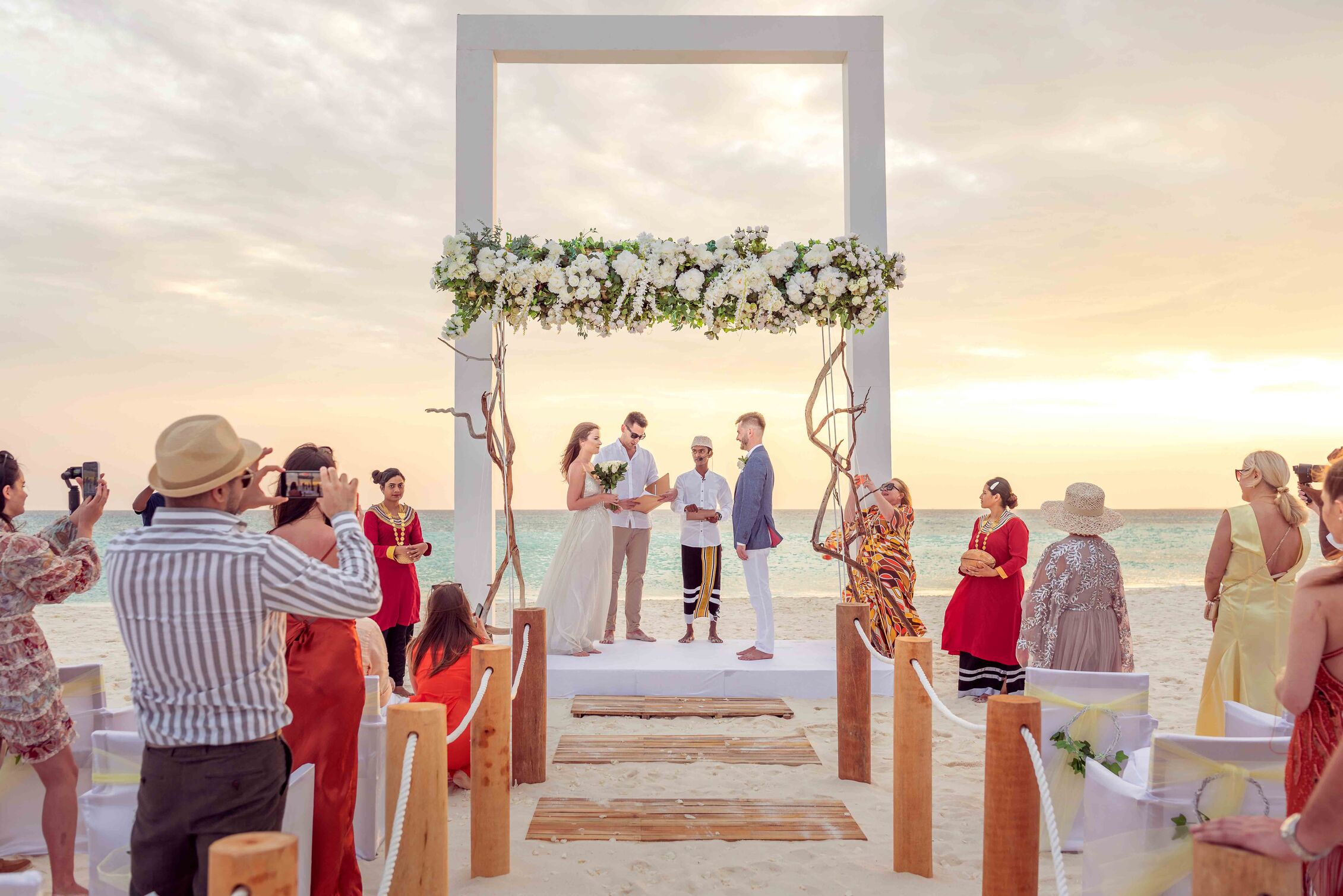 Bride and groom exchange vows beneath a white floral arch on a sunny beach, with guests taking photos and officiant nearby at sunset.