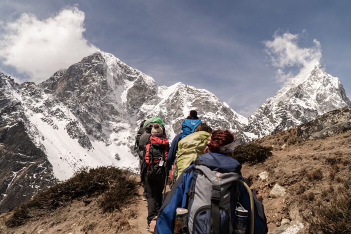 Hikers with backpacks climbing a mountain trail toward snow-covered peaks in the distance.