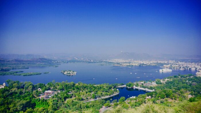 Aerial landscape of a large lake with small green islands, trees in the foreground, and distant mountains under a clear blue sky.