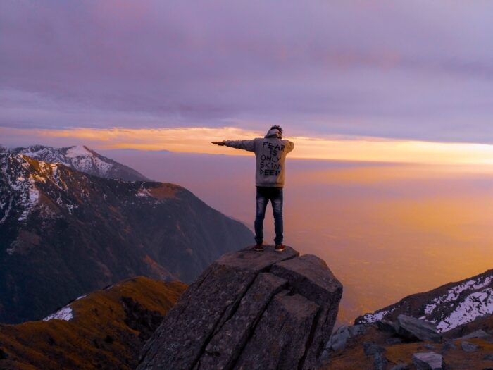 Person standing on a rocky cliff with arms outstretched at sunset, overlooking snow-capped mountains below.
